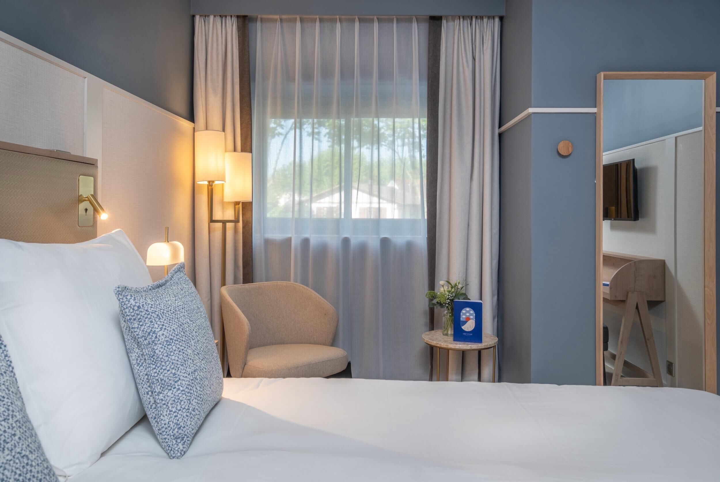 Habitación de hotel con cama, una silla, una mesita con un florero y un televisor sobre un pedestal.