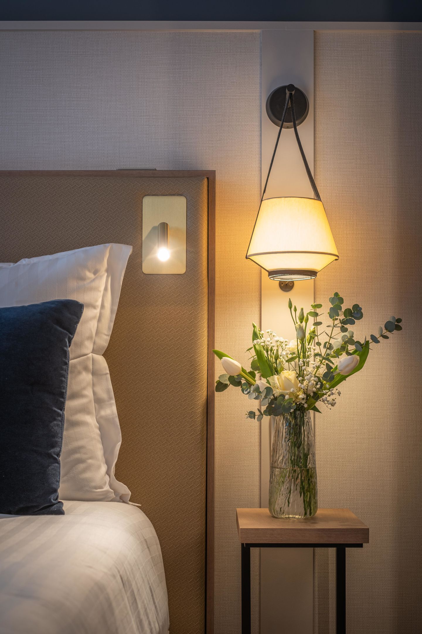 Una habitación de hotel con una cama, una mesita de noche con una lámpara y un florero con flores.