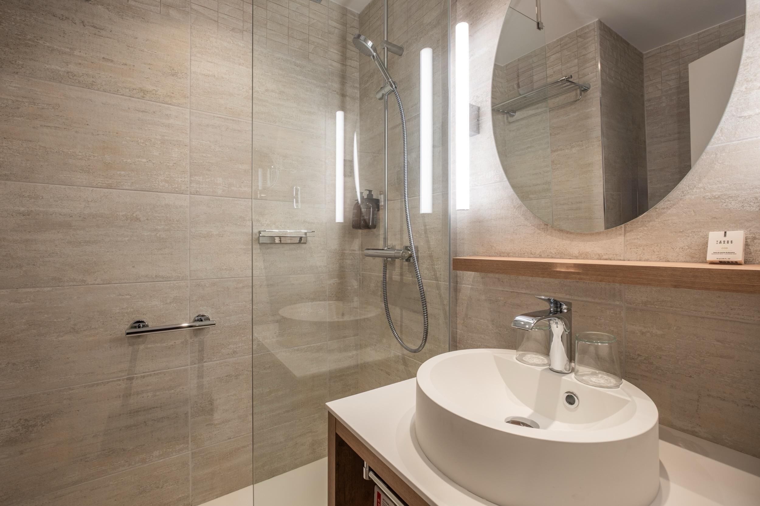 Un baño moderno con ducha envuelta en vidrio, un espejo redondo y un lavamanos blanco.