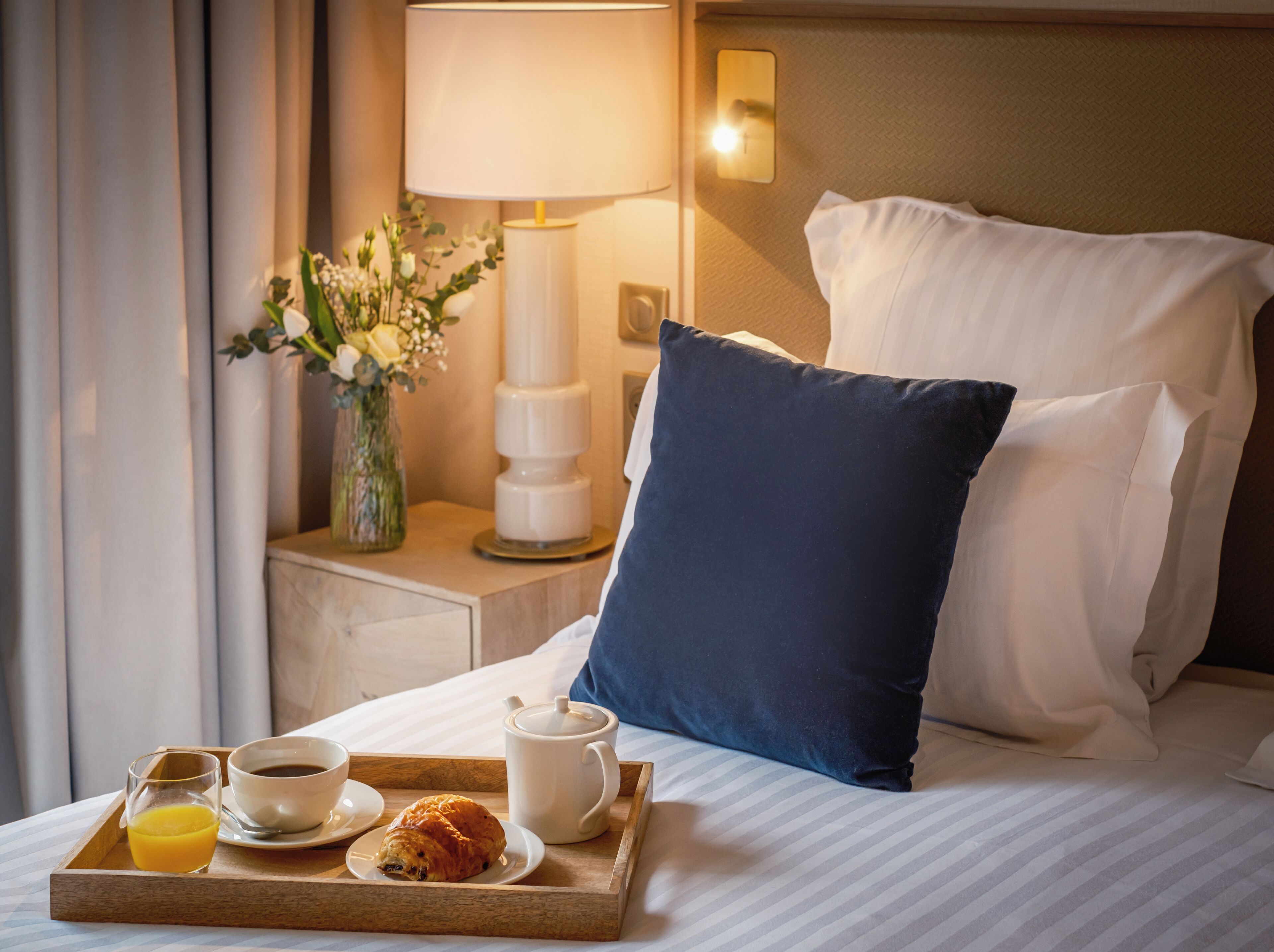 A hotel room with a neatly made bed, a bedside table with a lamp, a vase with flowers, and a tray with breakfast items including a croissant, coffee, and orange juice.