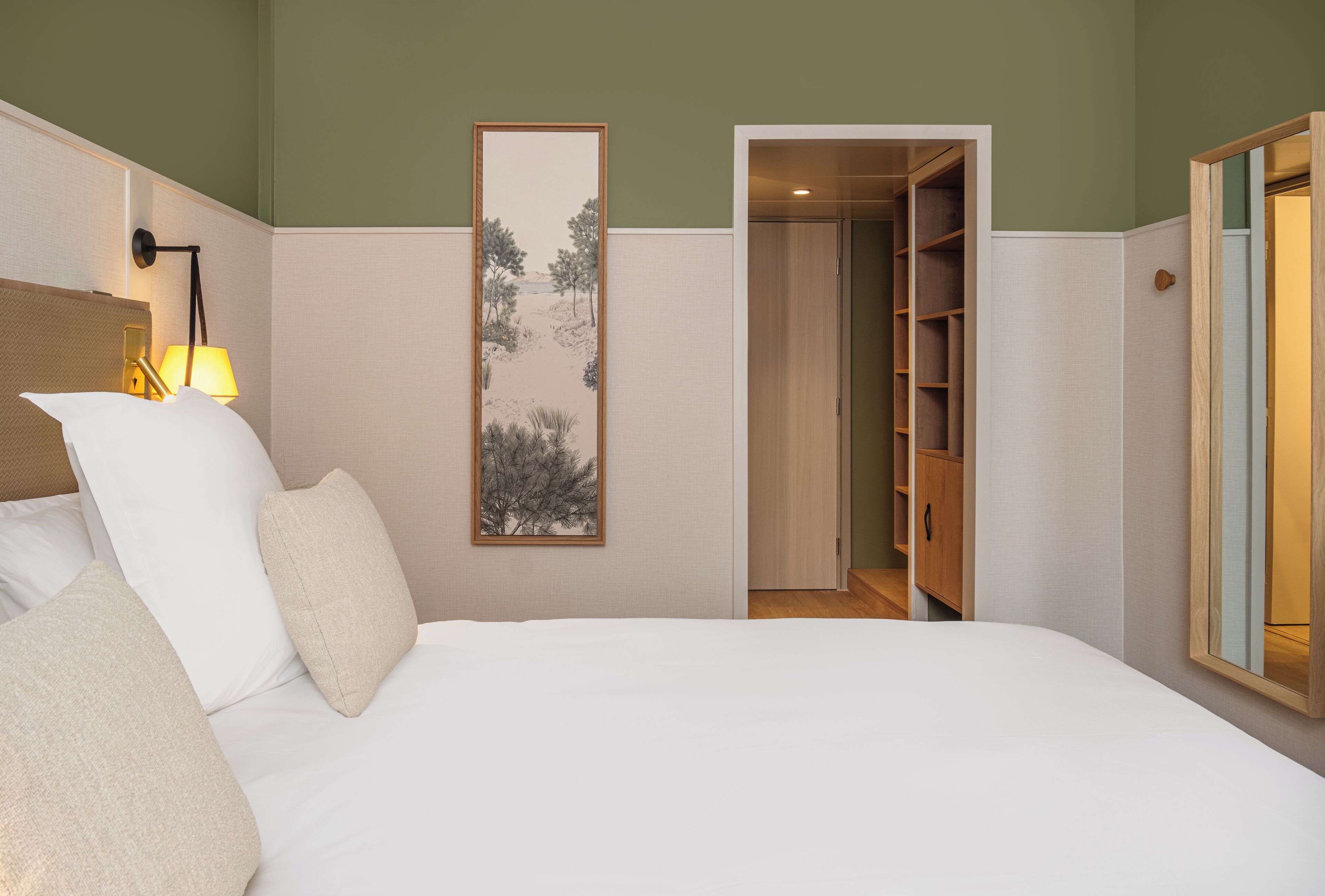 Una habitación de hotel con cama, mesita de noche con lámpara, un cuadro en la pared y un armario con espejo.
