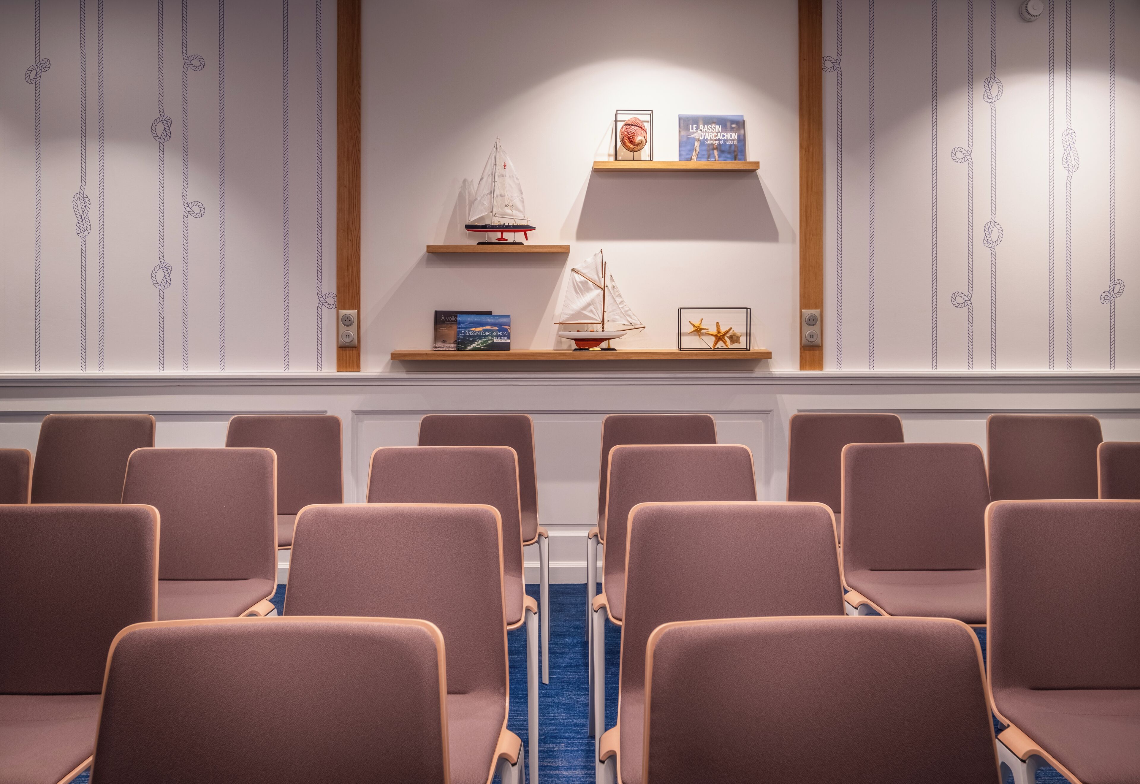 A conference room with rows of chairs, a blue carpet, and shelves with books and model ships.