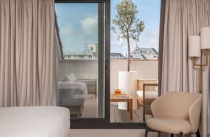 A hotel room with a bed, a chair, a table with a teapot and glass, and a view of buildings and a tree through a glass door. - Arcanse by Inwood Hotels (Arcachon)