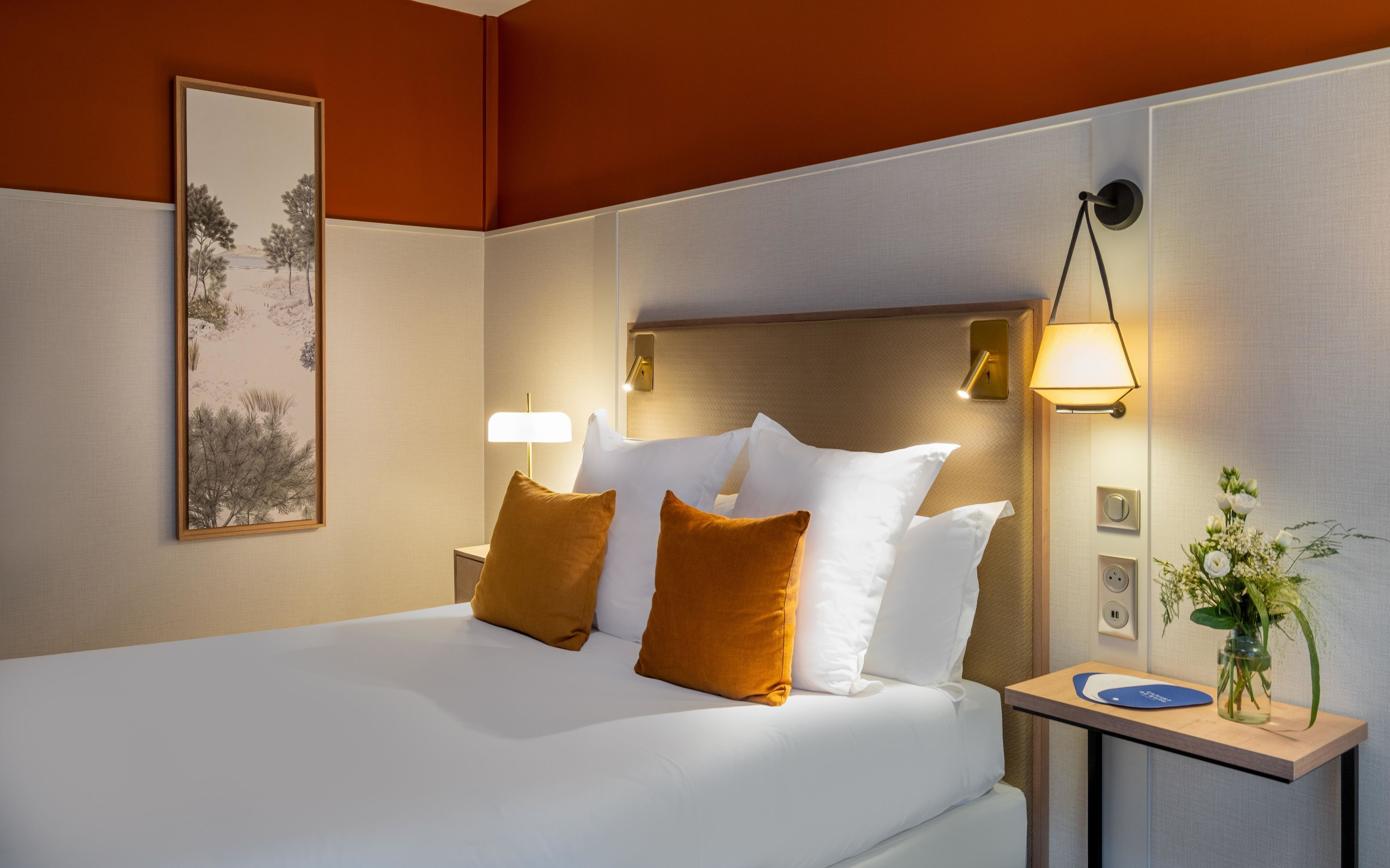 A hotel room with a bed, pillows, a nightstand, a lamp, a wall clock, and a framed picture.