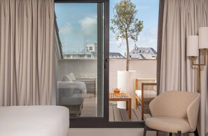 A hotel room with a bed, a chair, a table with a lamp, and a view of buildings and a tree through a glass door. - Arcanse by Inwood Hotels (Arcachon)