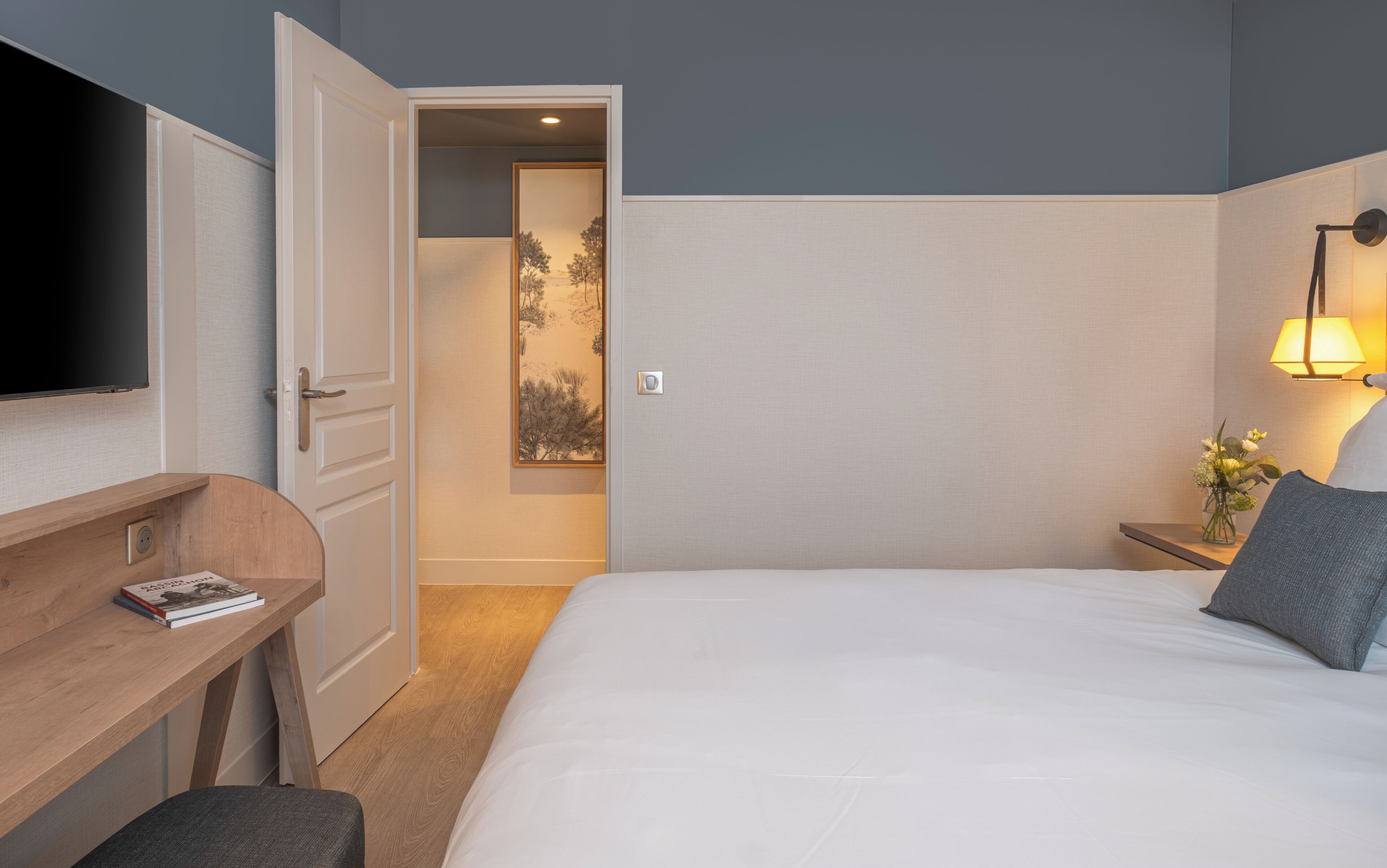 A hotel room with a bed, a desk, a chair, a TV, a bedside lamp, and a small table with a vase of flowers.