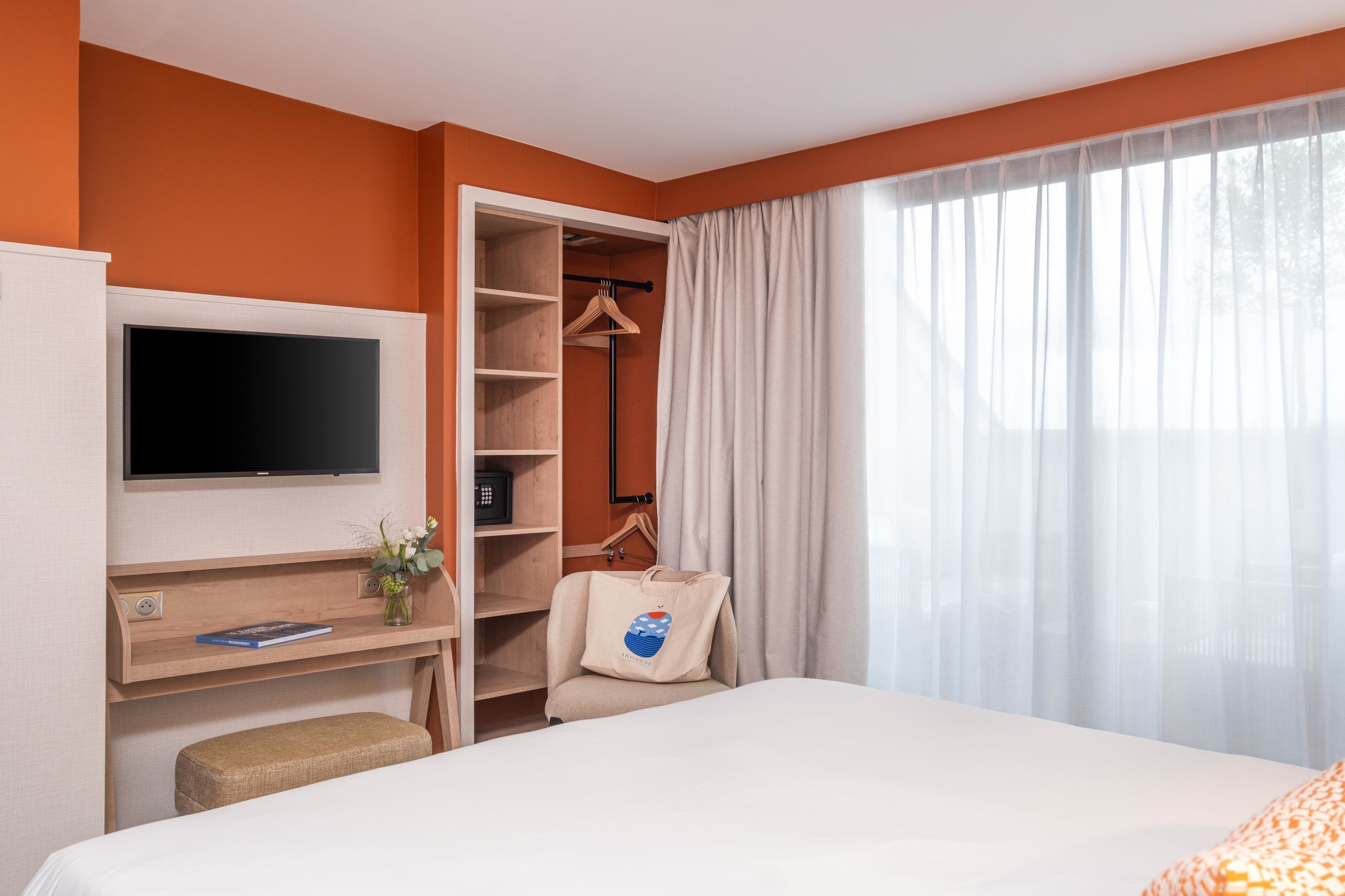Quarto de hotel com cama, televisão, secretária com cadeira, guarda-roupa e janela com cortinas.