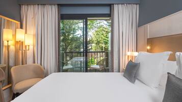 Habitación de hotel con una cama grande, dos sillones, un balcón con mesa y sillas, y dos lámparas fijadas a la pared.