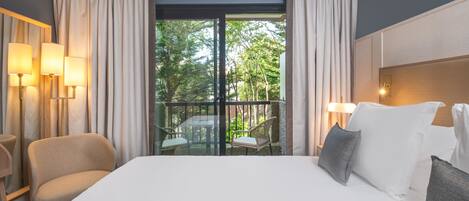 Habitación de hotel con una cama grande, dos sillones, un balcón con mesa y sillas, y dos lámparas fijadas a la pared.