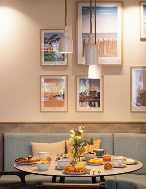 A dining area with a round table set for breakfast, a sofa, and framed artwork on the wall. - Arcanse by Inwood Hotels (Arcachon)