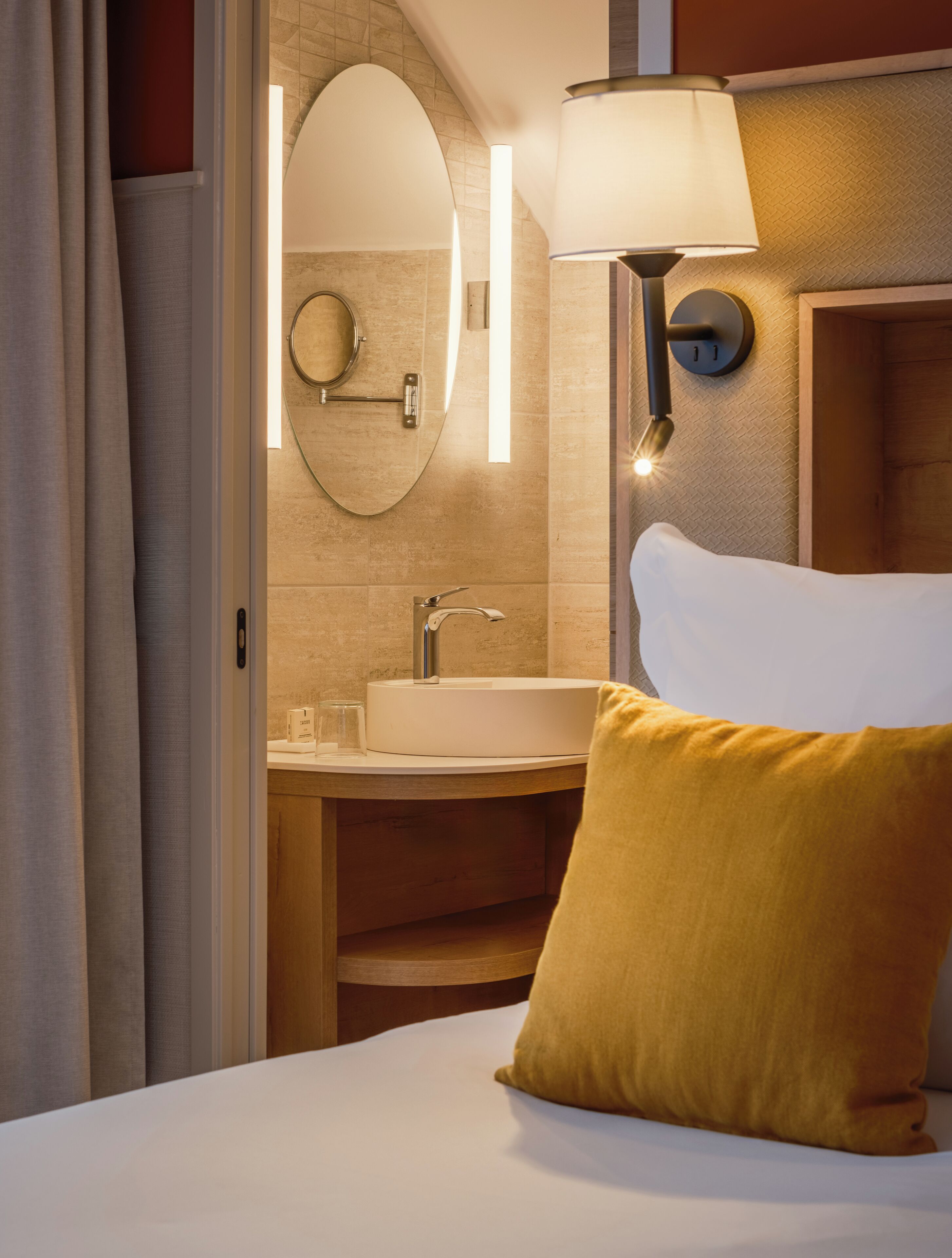 A hotel room with a bed, a bedside lamp, a round mirror, a sink, and a wooden headboard.