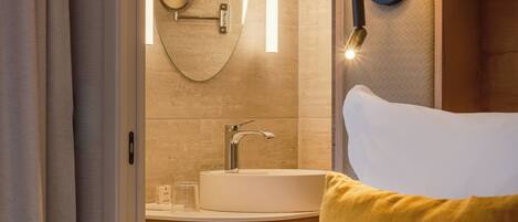 A hotel room with a bed, a bedside lamp, a round mirror, a sink, and a wooden headboard.
