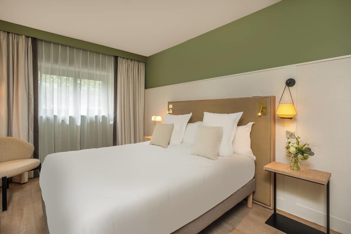 A hotel room with a large bed, a bedside table with a lamp, a chair, and a window with curtains.
