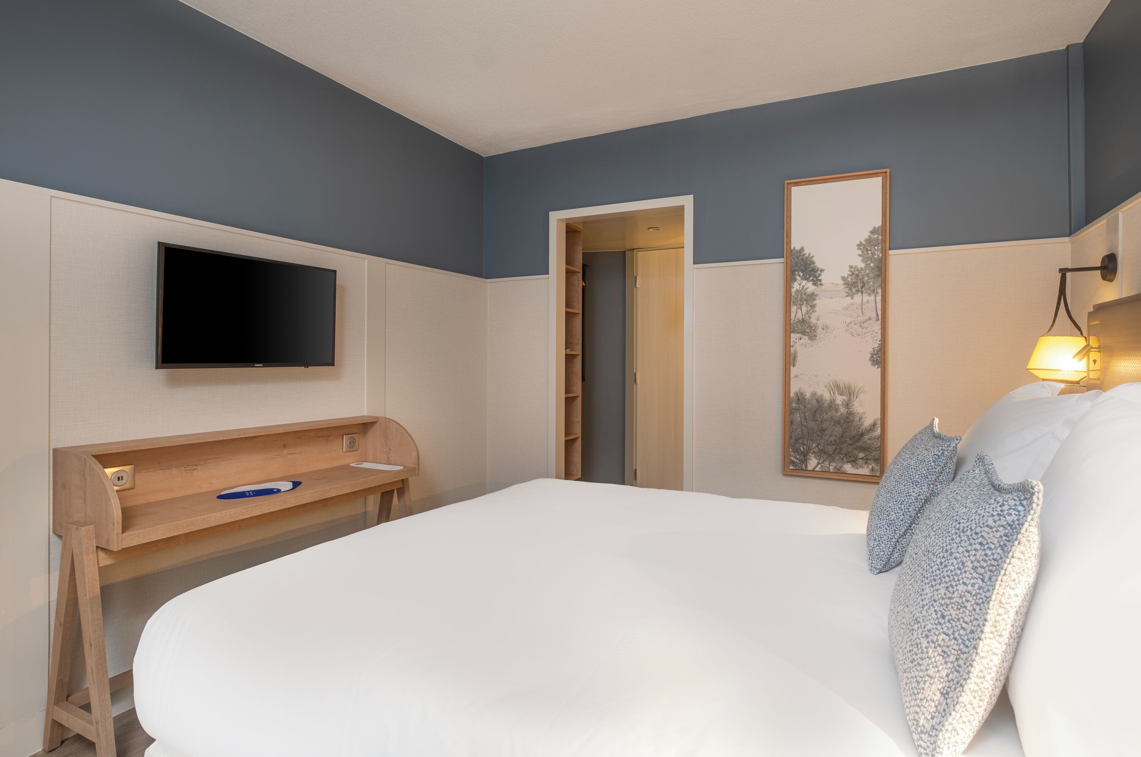 Ein Hotelzimmer mit einem großen Bett, einem Schreibtisch mit Stuhl, einem an der Wand befestigten Flachbildfernseher, einer zur Garderobe führenden Tür und einem gerahmten Bild an der Wand.