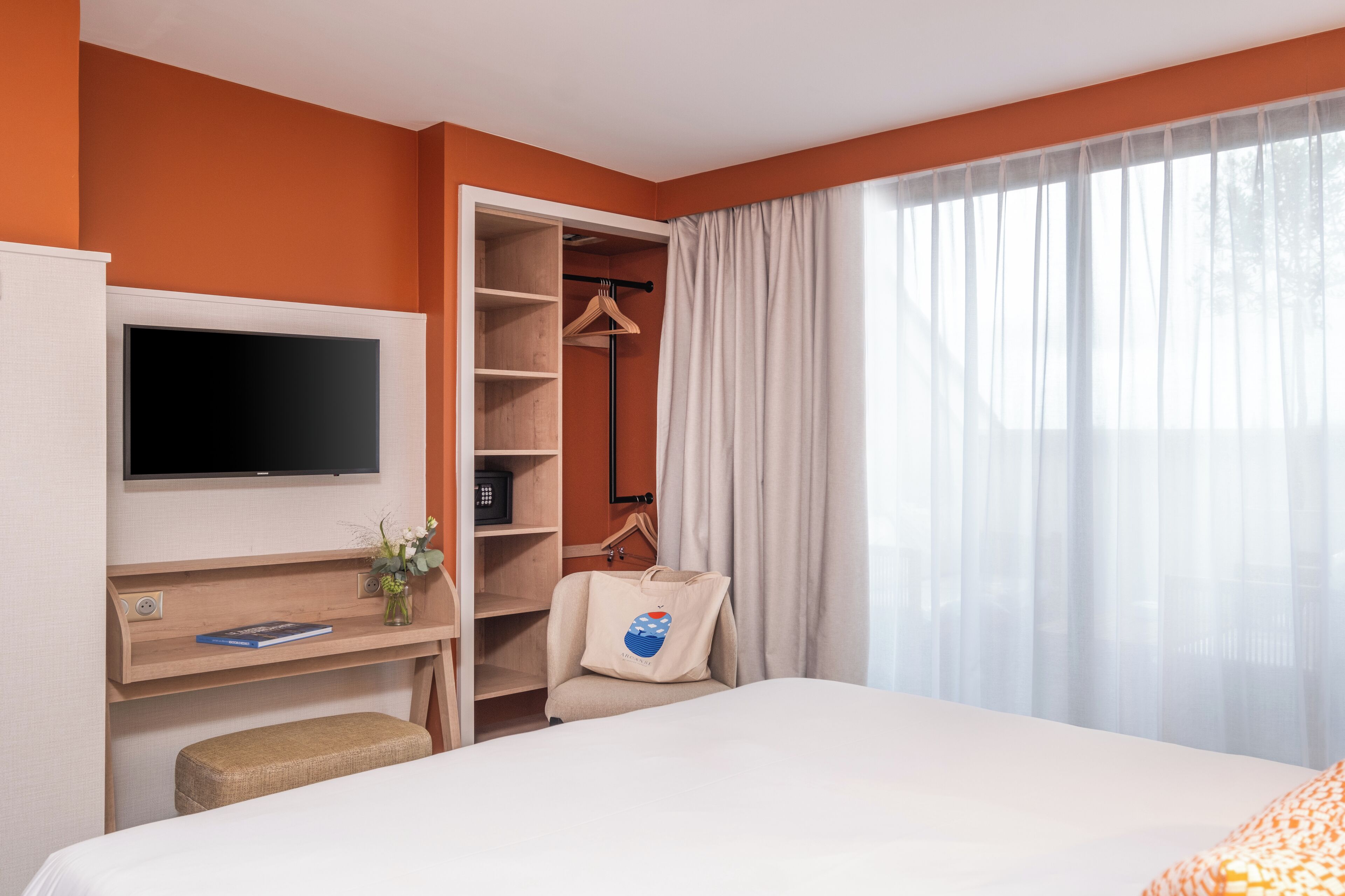 A hotel room with a bed, a TV mounted on the wall, a desk with a computer, a chair, a wardrobe, and a window with curtains.