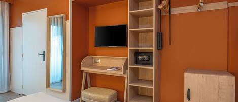 Una habitación de hotel con cama, escritorio con computadora, televisor, armario y un taburete pequeño.