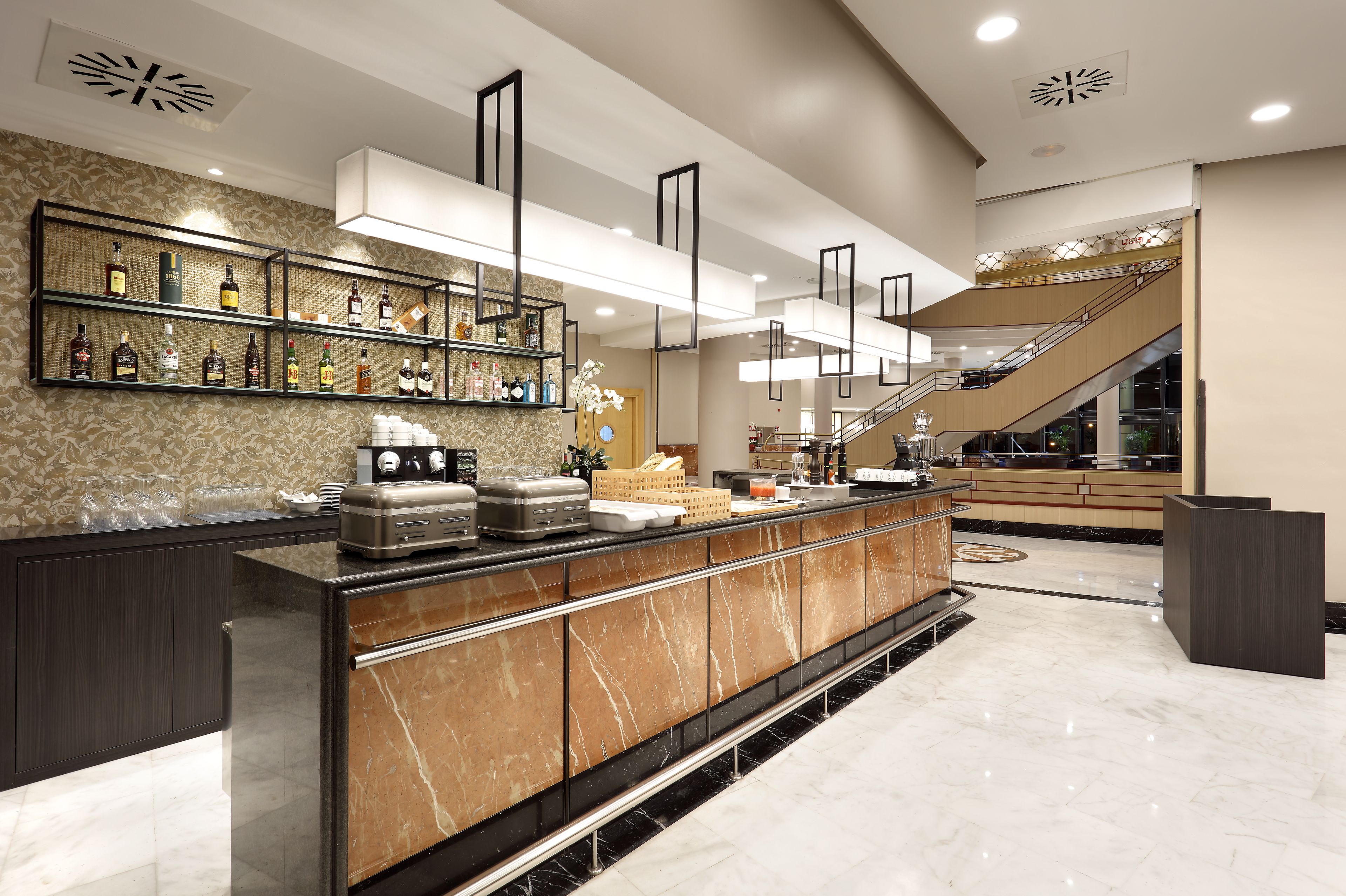 A modern hotel lobby with a marble reception desk, a variety of liquor bottles on shelves, a coffee station, and a staircase leading to an upper level.