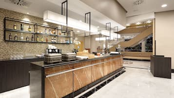 A modern hotel lobby with a marble reception desk, a variety of liquor bottles on shelves, a coffee station, and a staircase leading to an upper level.
