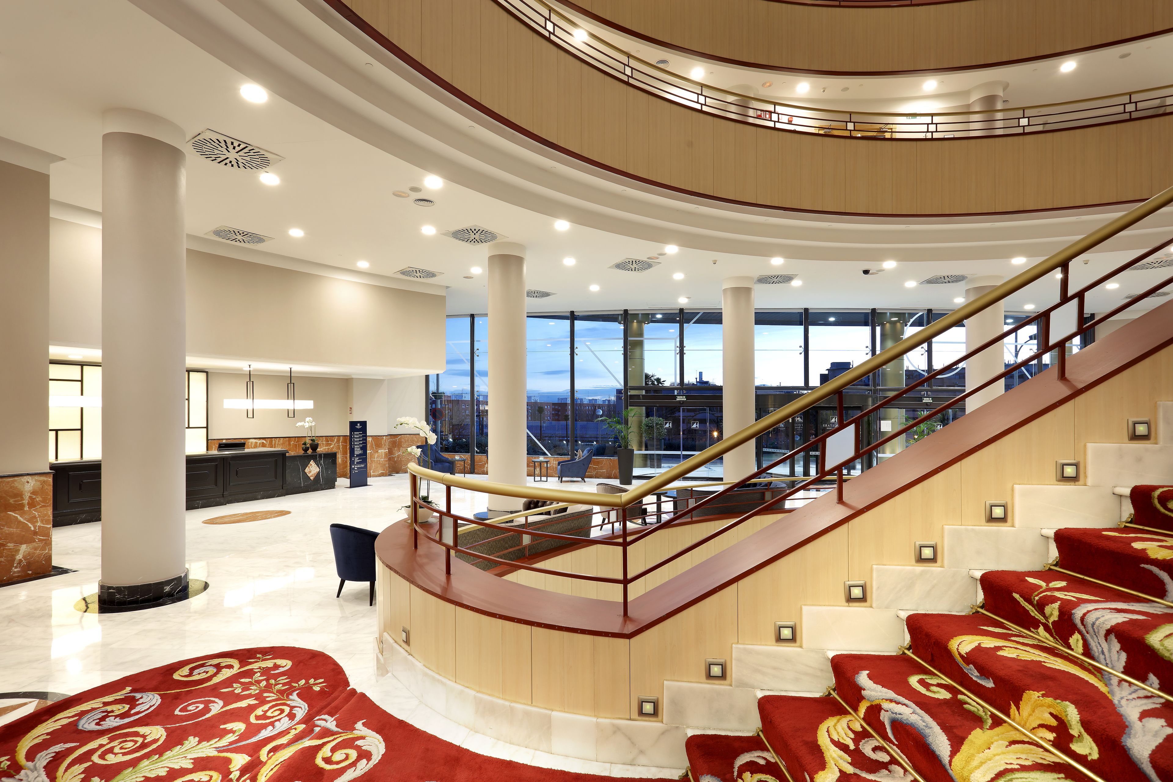 A spacious lobby with a curved staircase, a reception desk, and a view of the cityscape through large windows.