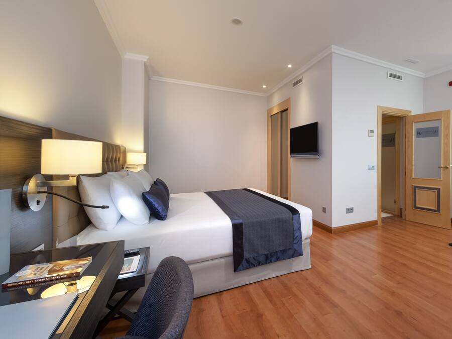 A hotel room with a bed, bedside table, lamp, TV, and a desk with a chair.