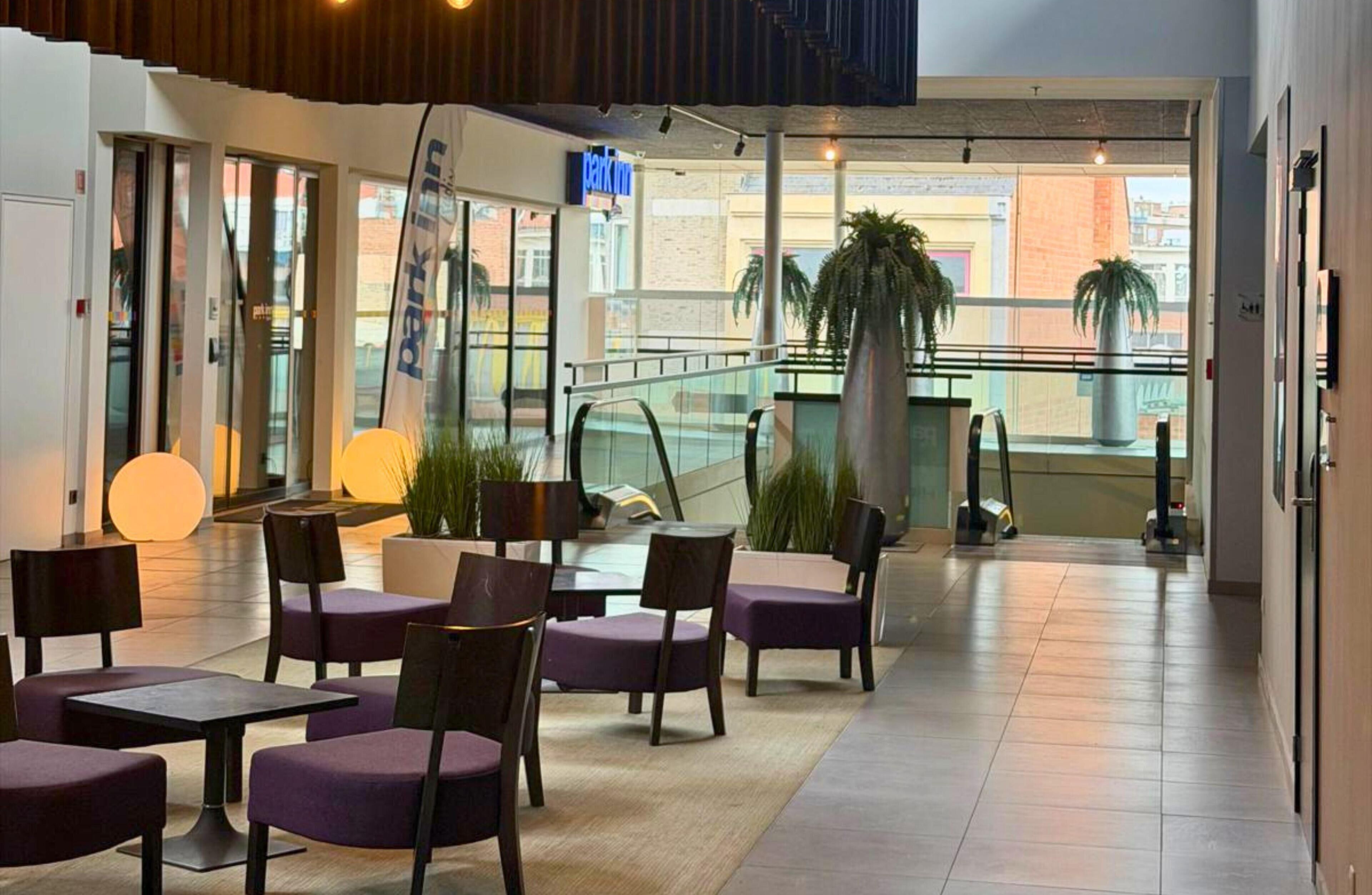 A modern indoor seating area with tables and chairs, a staircase, and large windows overlooking a cityscape.