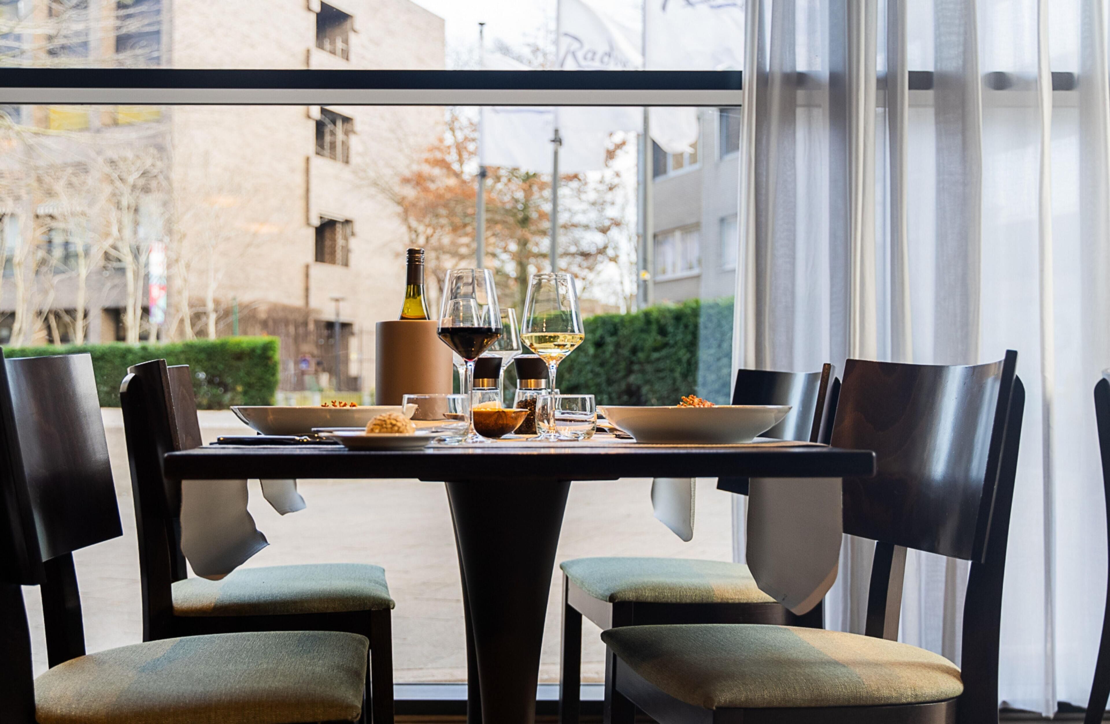 Een eettafel met stoelen, een wijnfles en glazen, met uitzicht op een gebouw en bomen.