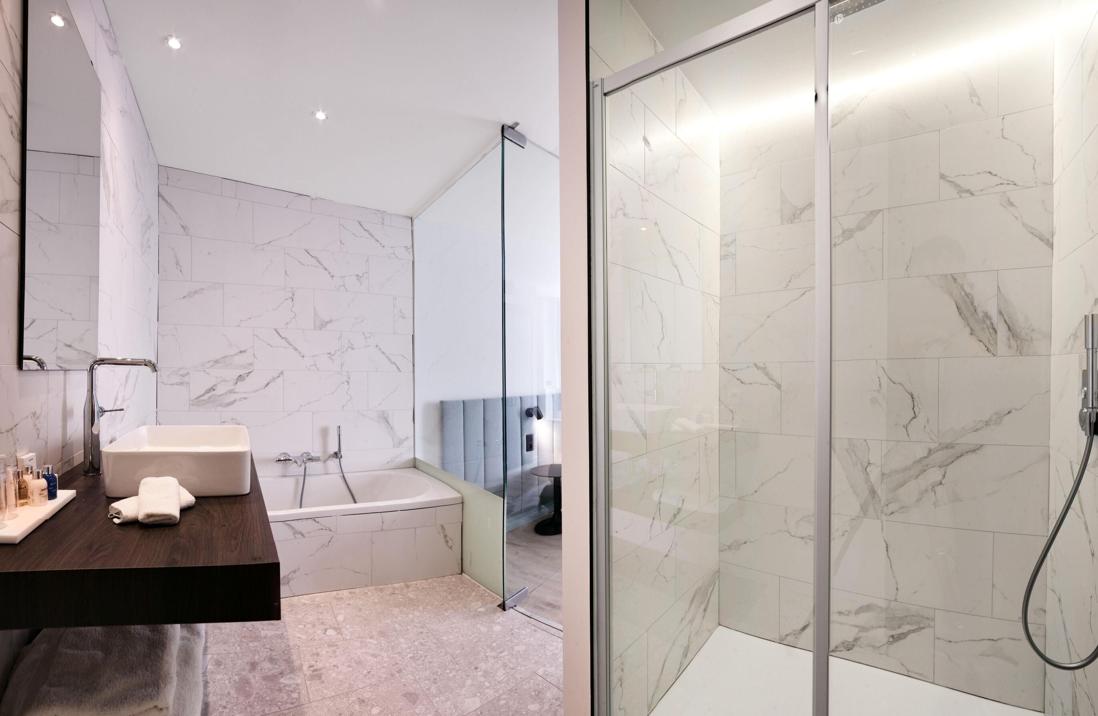 Un baño moderno con pared de azulejos de mármol, una bañera independiente, ducha con cerramiento de vidrio y un mueble de tocador de madera oscura.