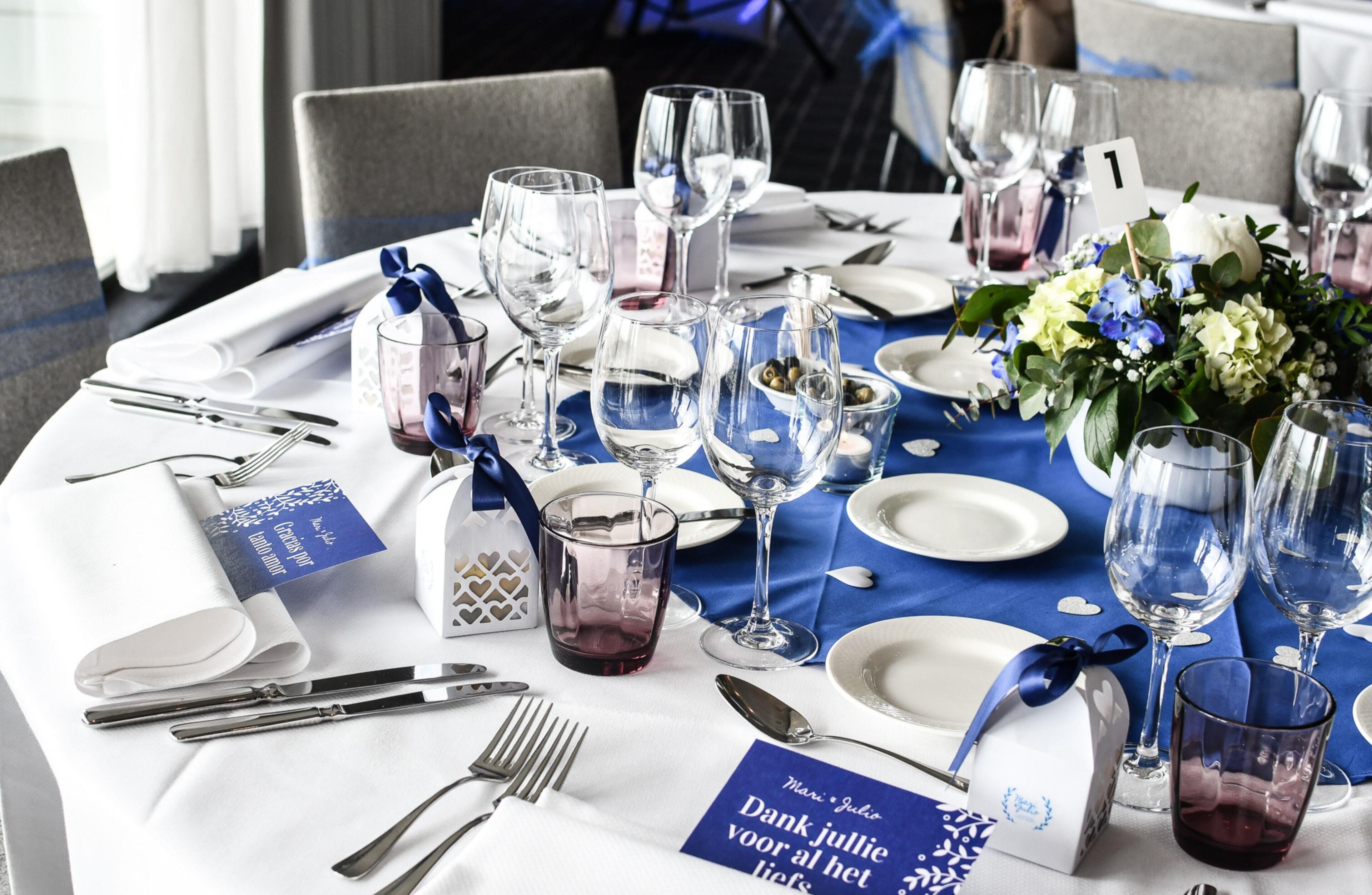 Una mesa dispuesta con copas de vino, platos, cubiertos y un mantel azul.