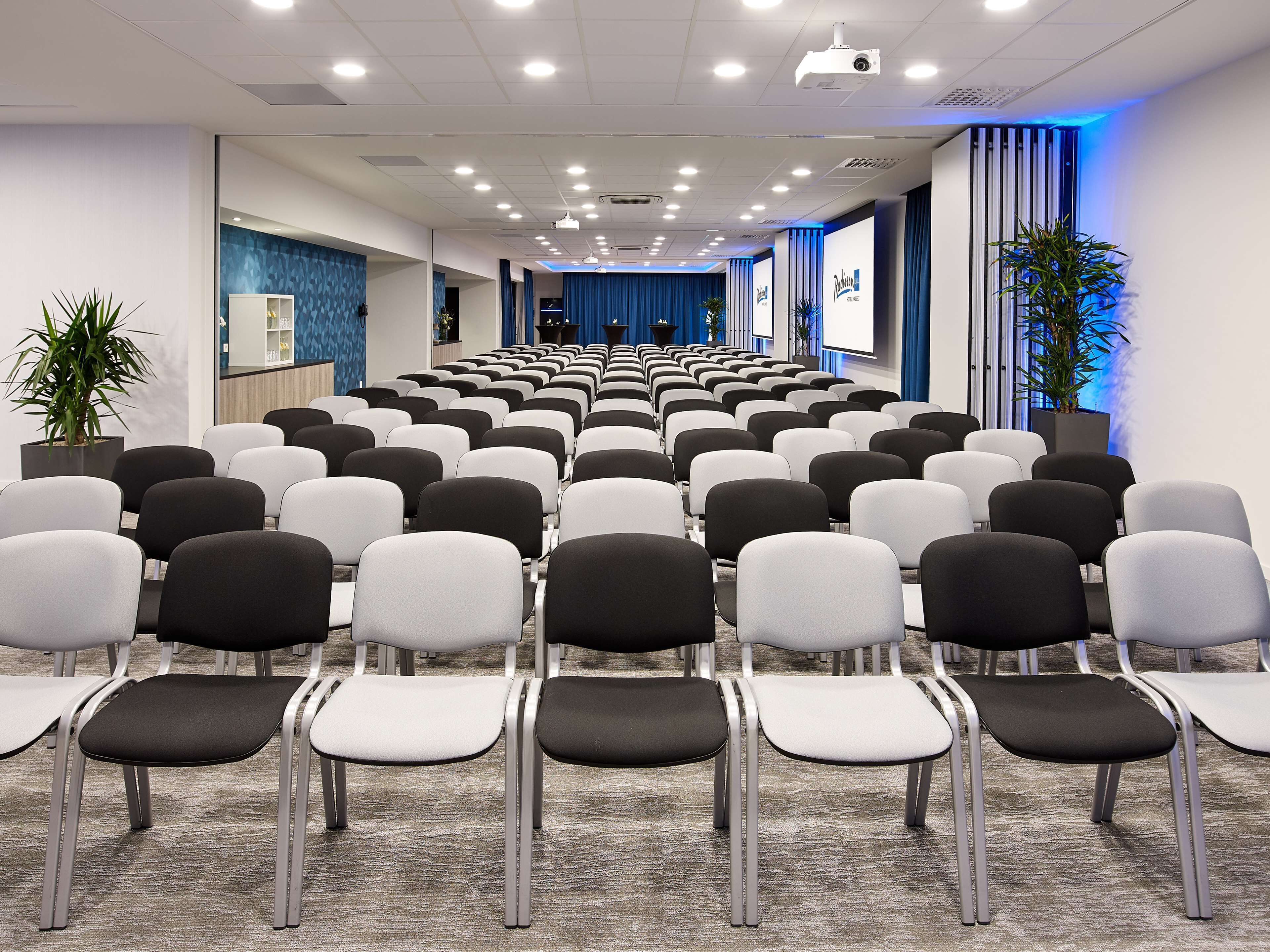 Ein Konferenzraum mit Stuhlreihen, einer Projektionsleinwand und einer blau beleuchteten Wand.