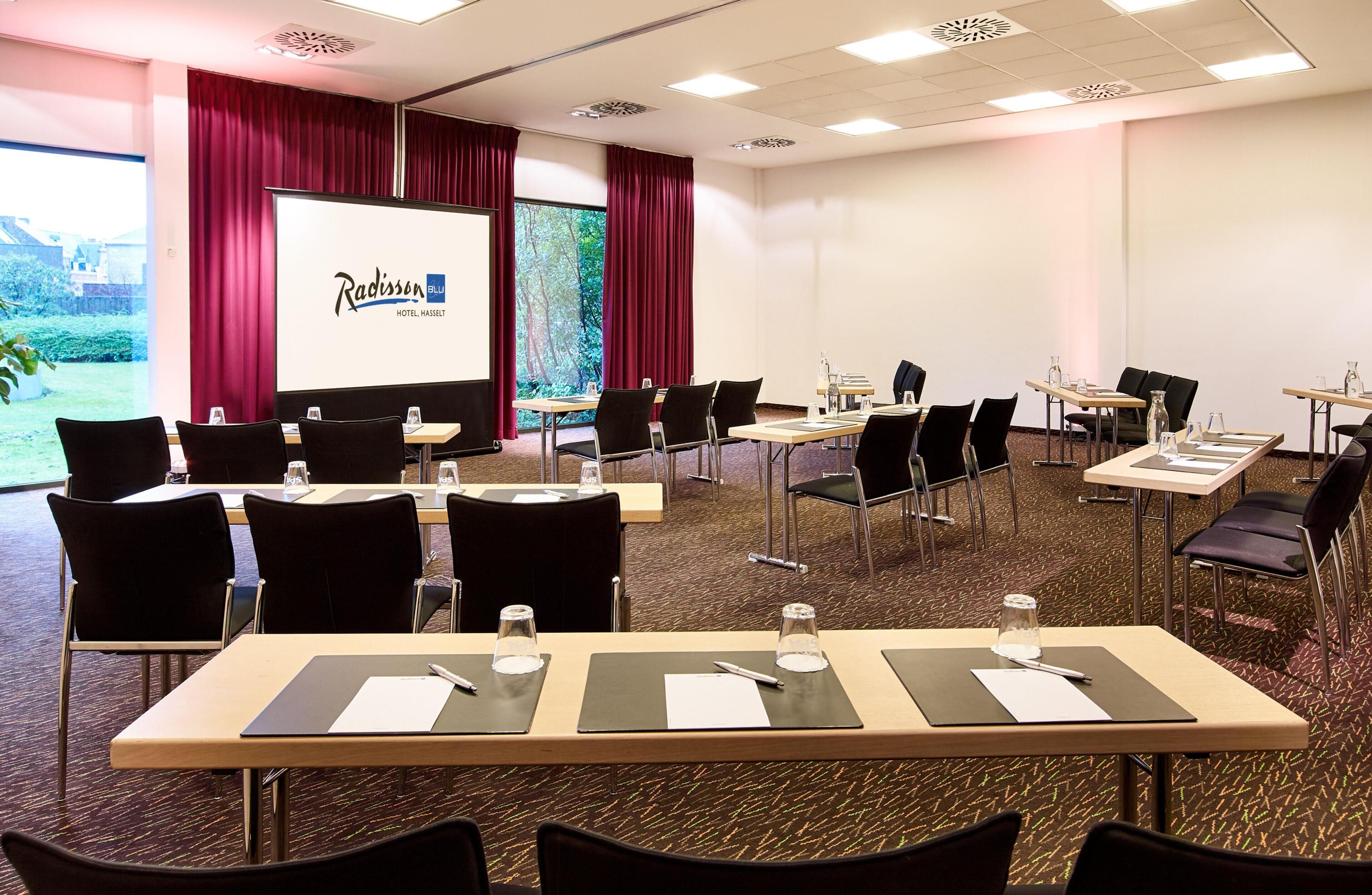 Un salón de conferencias con el letrero de un hotel Radisson Blu, varias mesas con sillas y una pantalla de proyector.