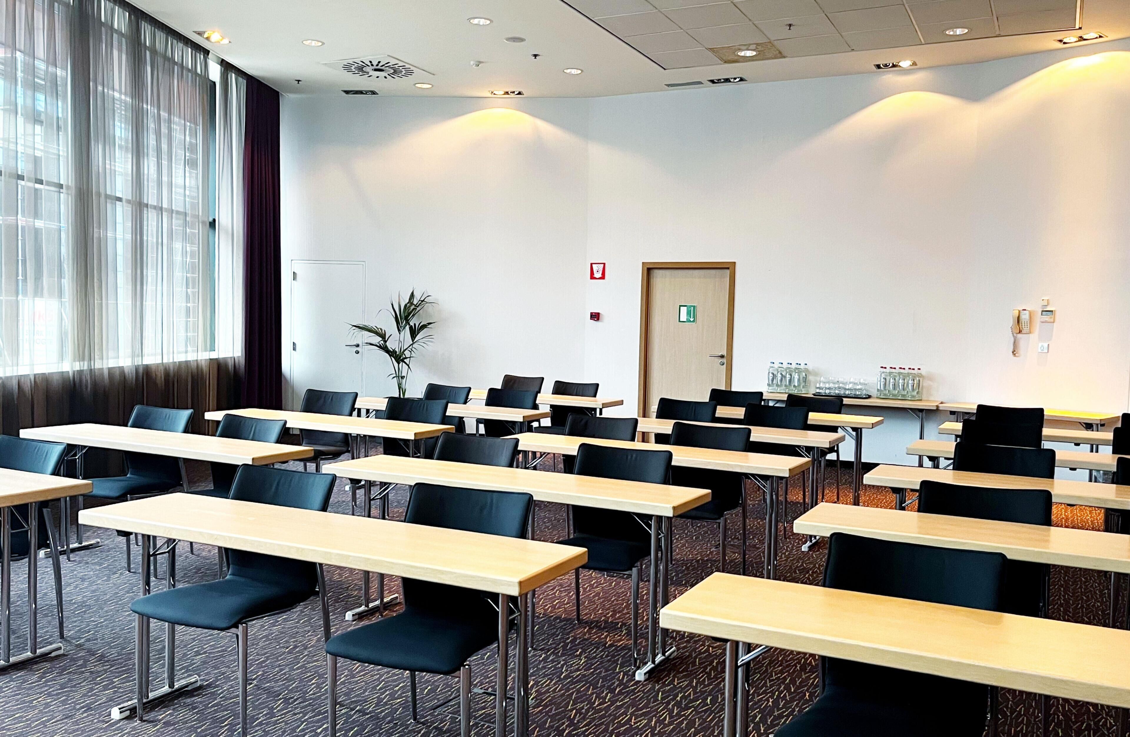 Ein Konferenzraum mit Reihen von Tischen und Stühlen, einer Tür mit einem Notausgangsschild und einer Pflanze in der Ecke.