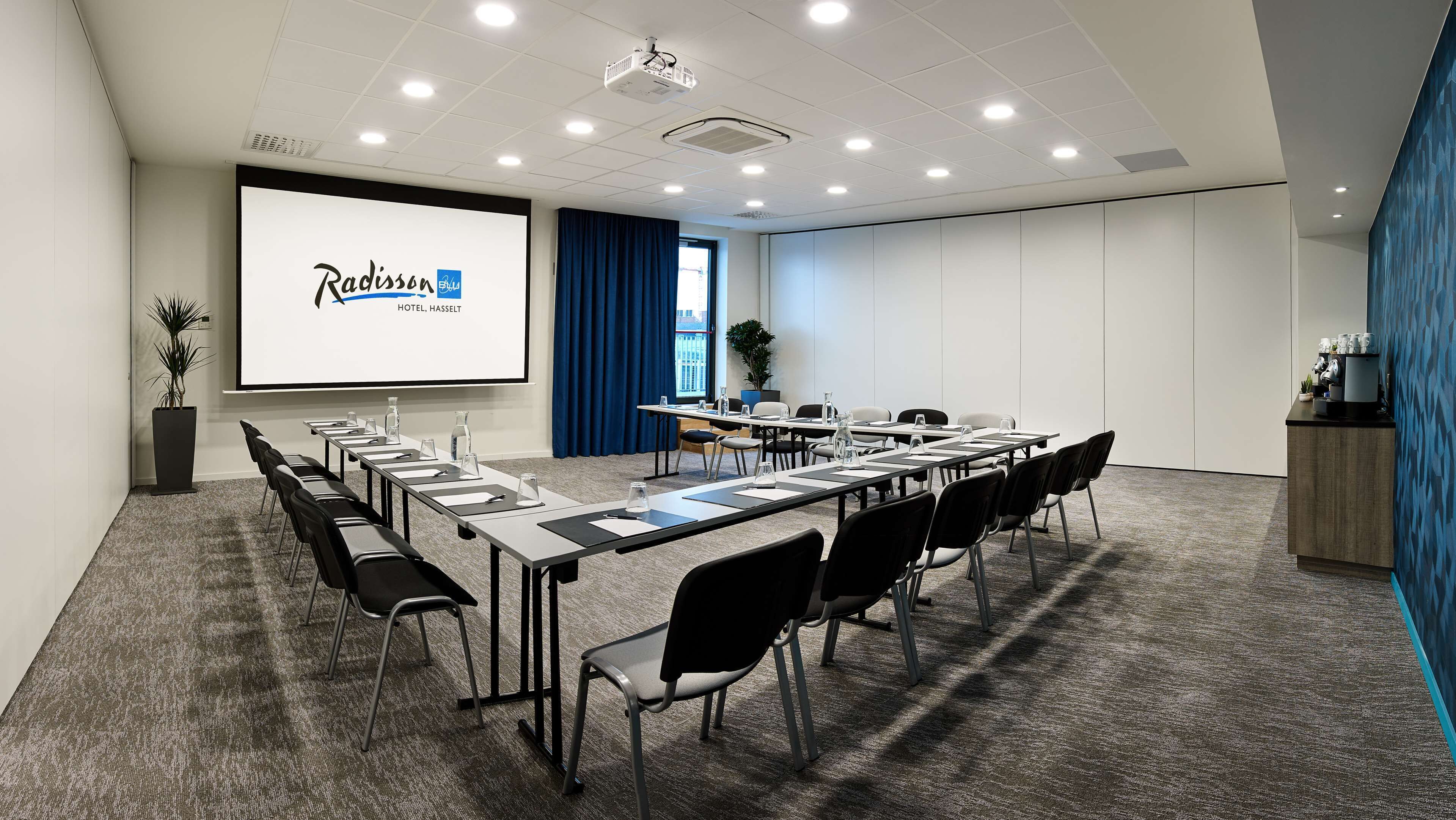 A conference room with a long table, chairs, a projector screen displaying Radisson Hotel logo, and a coffee station.