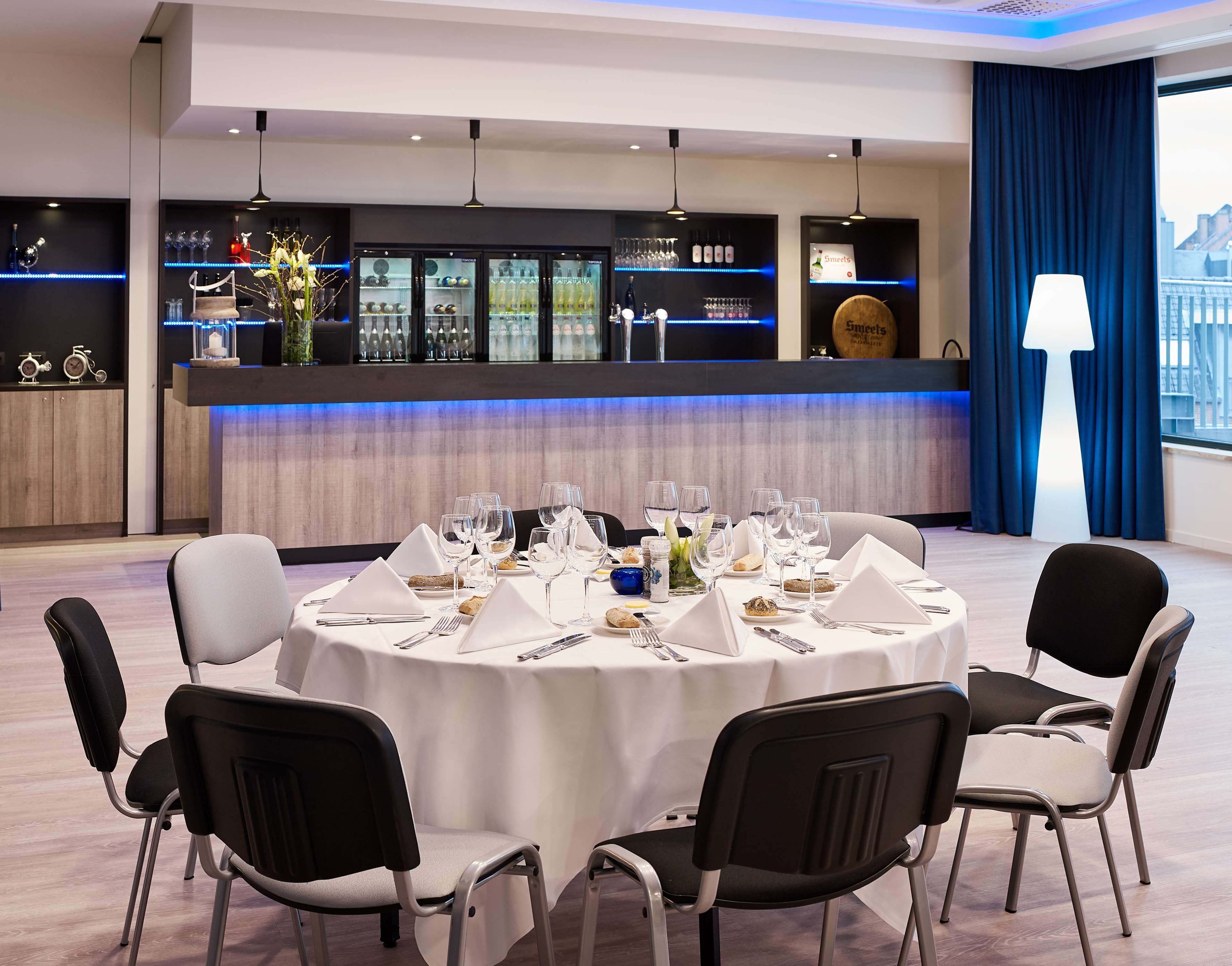 A round dining table set for six with chairs, a bar area with shelves of glasses and bottles, and a view of the city through large windows.