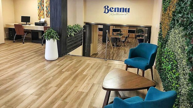 Uma área de recepção com piso de madeira, um balcão com computador, uma planta em vaso e uma parede com o letreiro 'Bennu'.