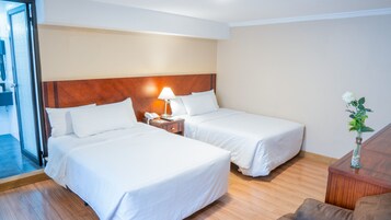 Habitación de hotel con dos camas, cabecera de madera, mesita de noche con lámpara y un jarrón con una flor.