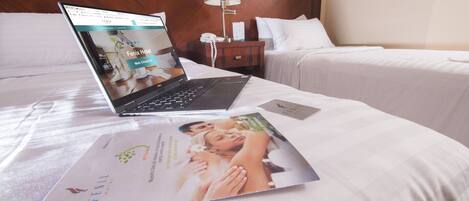 A hotel room with a laptop on the bed, a brochure, a nightstand with a lamp, and a bed with white bedding.