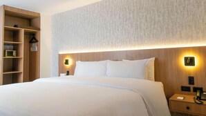 Quarto de hotel com uma cama grande, uma mesa de cabeceira de madeira, uma luminária fixada na parede e uma estante embutida.