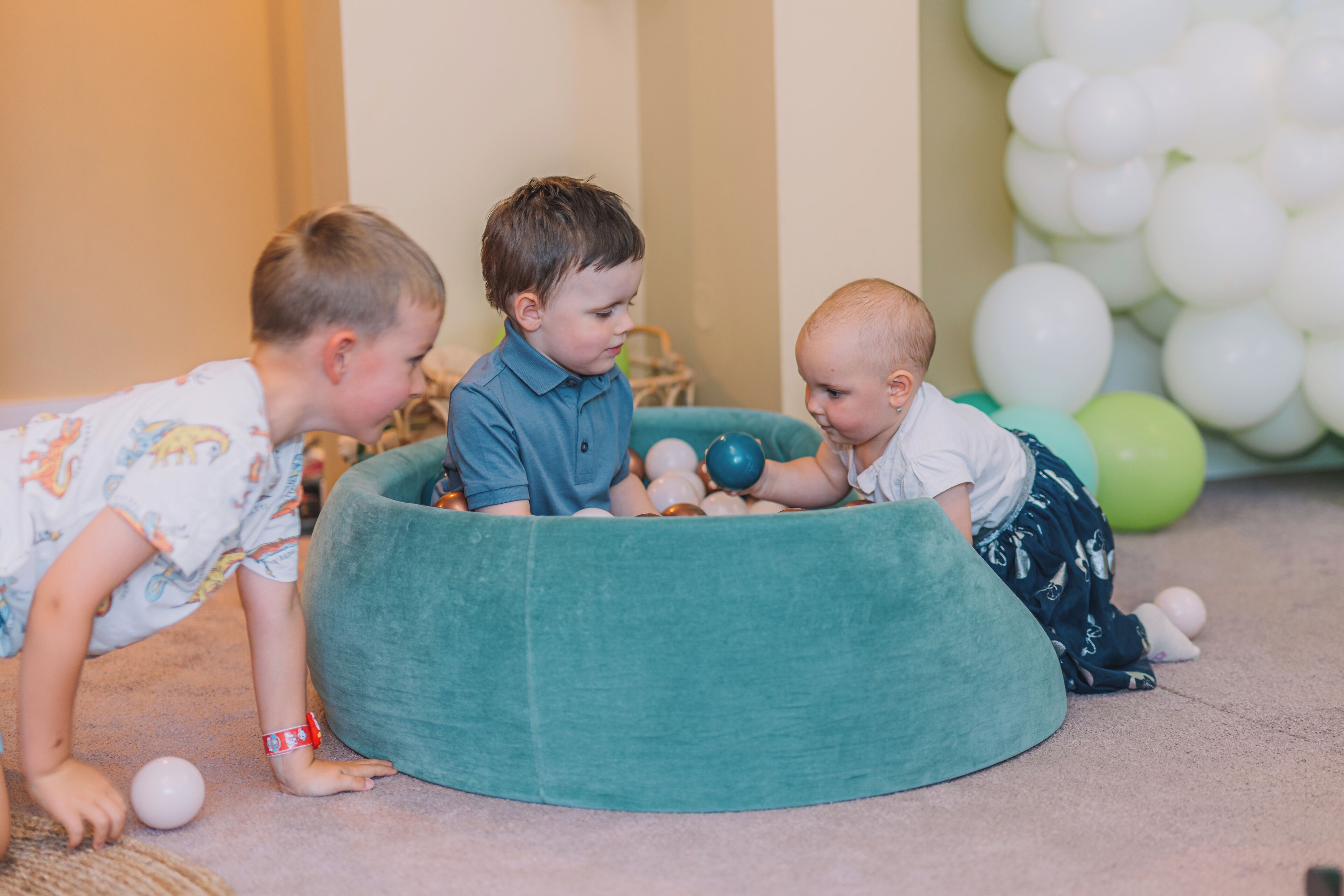 Trois enfants jouent avec des balles dans une pièce où se trouvent un fauteuil en forme de haricot vert et, à l’arrière-plan, des ballons.