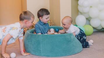 Drie kinderen spelen met ballen in een kamer met een groene relaxstoel en ballonnen op de achtergrond.