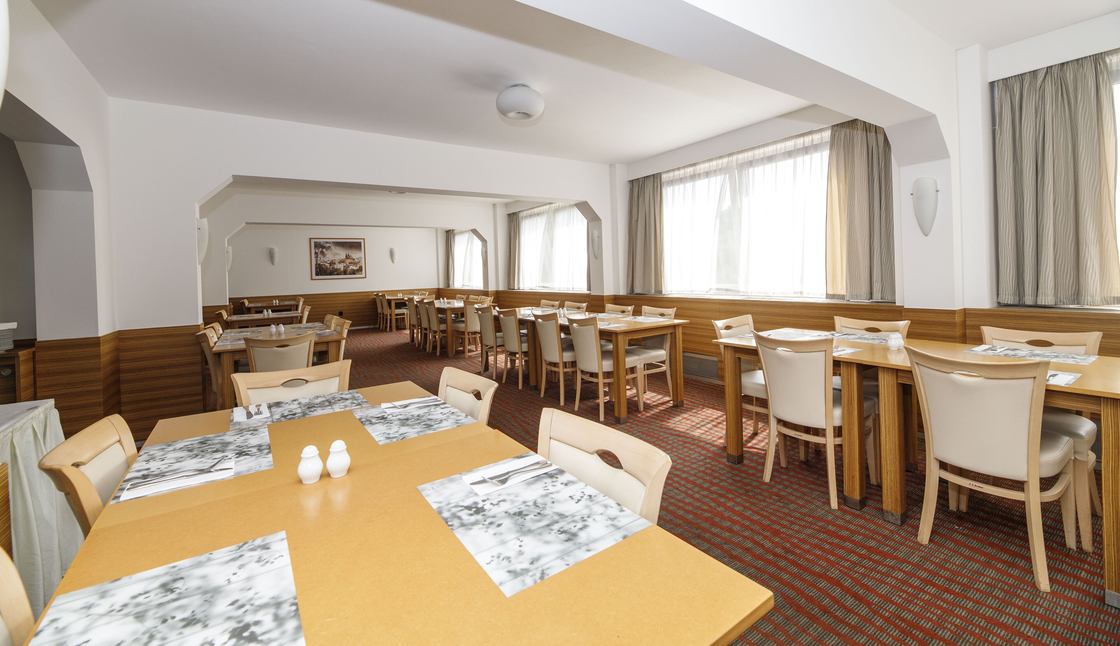 Un comedor de hotel con varias mesas y sillas, piso alfombrado de color rojo y grandes ventanales con cortinas.