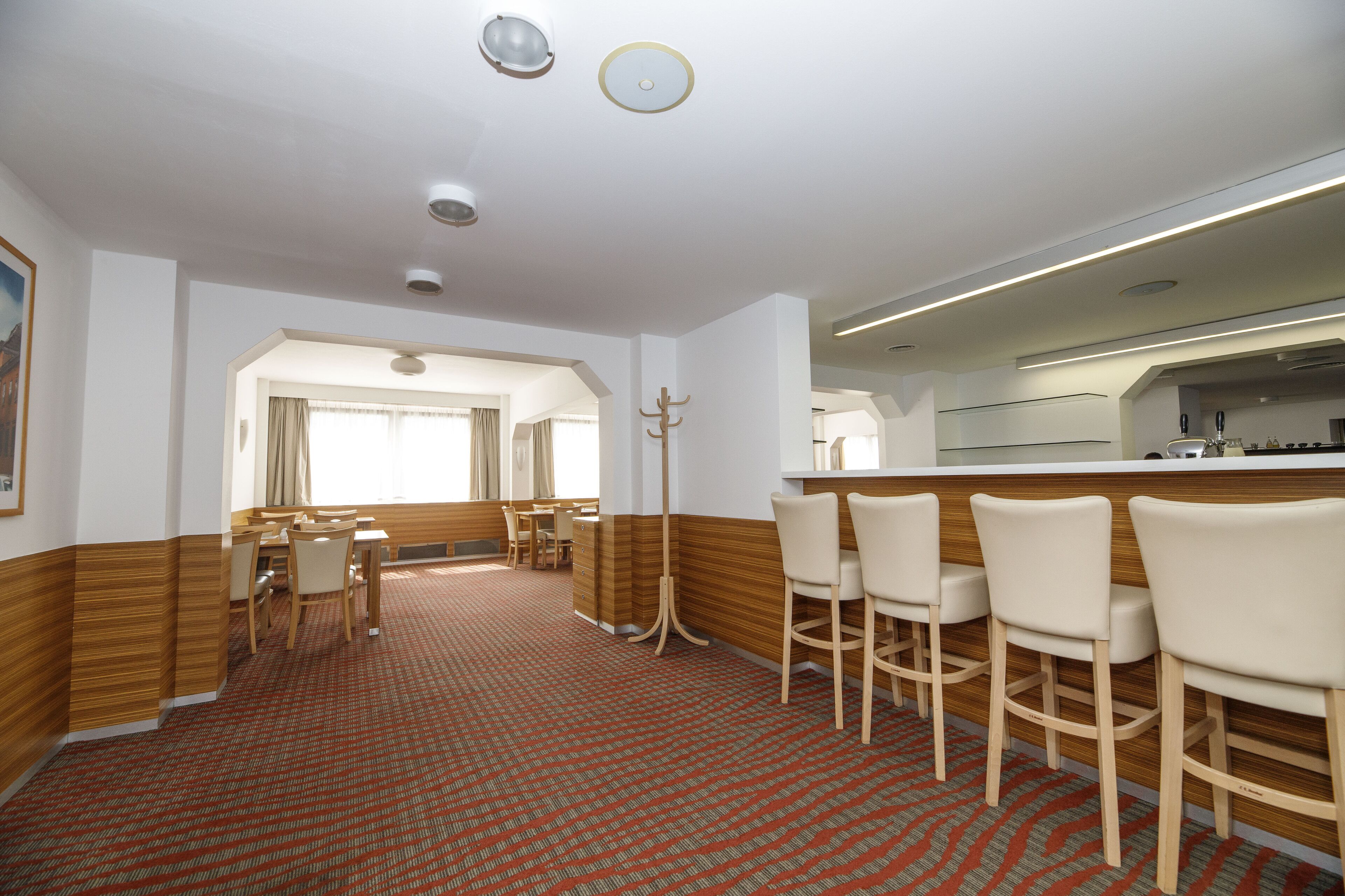 Un vestíbulo de hotel con alfombra roja estampada, detalles de madera, un mostrador de recepción y un área de comedor con sillas y mesas.