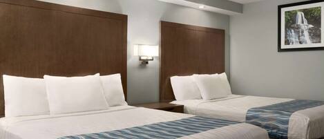 A hotel room with two beds, each with white bedding and a blue patterned throw. There is a wall-mounted lamp between the beds and a framed picture of a waterfall on the wall.