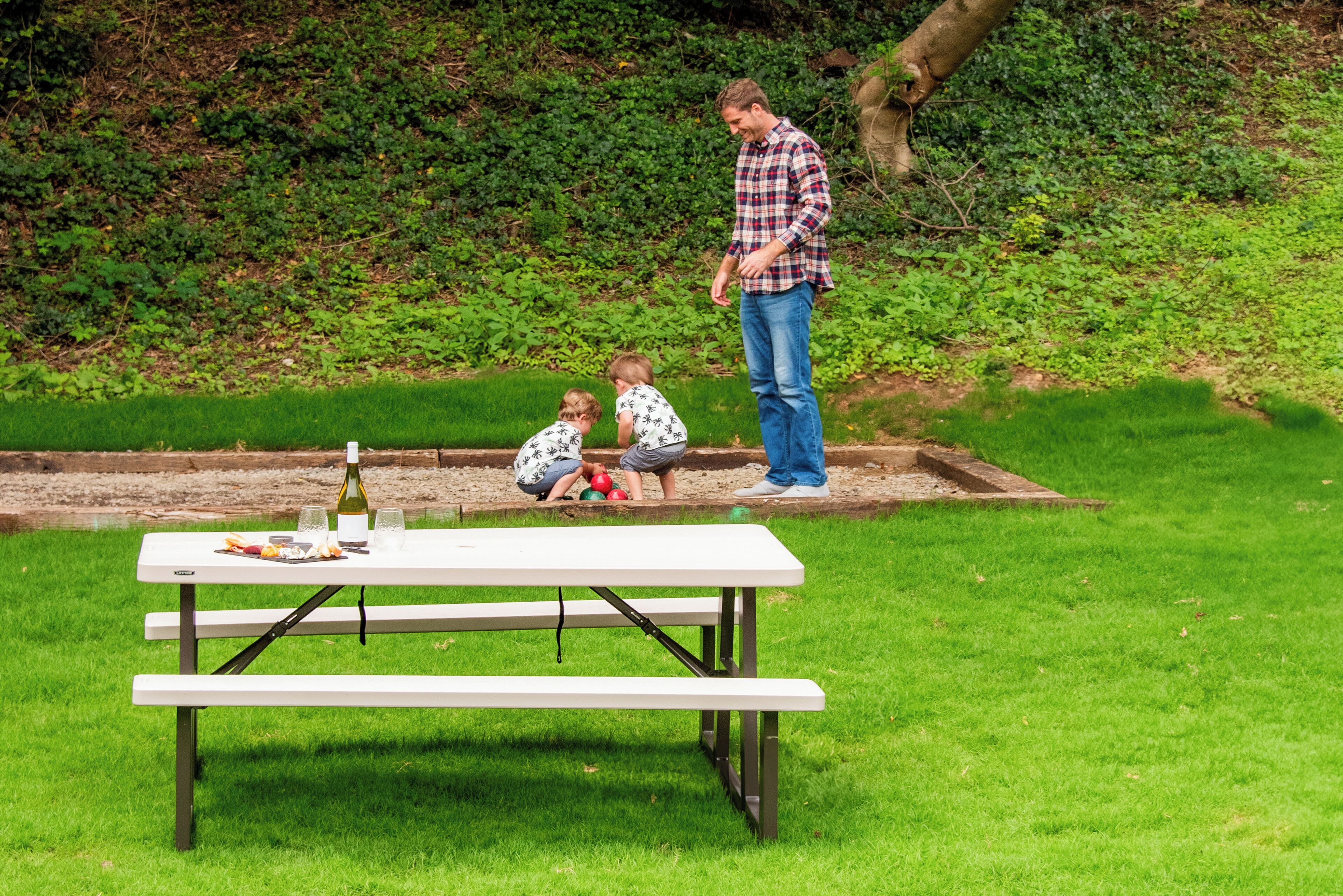 A picnic table with a bottle of wine and glasses, set on a grassy area with a tree and a hedge in the background.