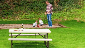 A picnic table with a bottle of wine and glasses, set on a grassy area with a tree and a hedge in the background.
