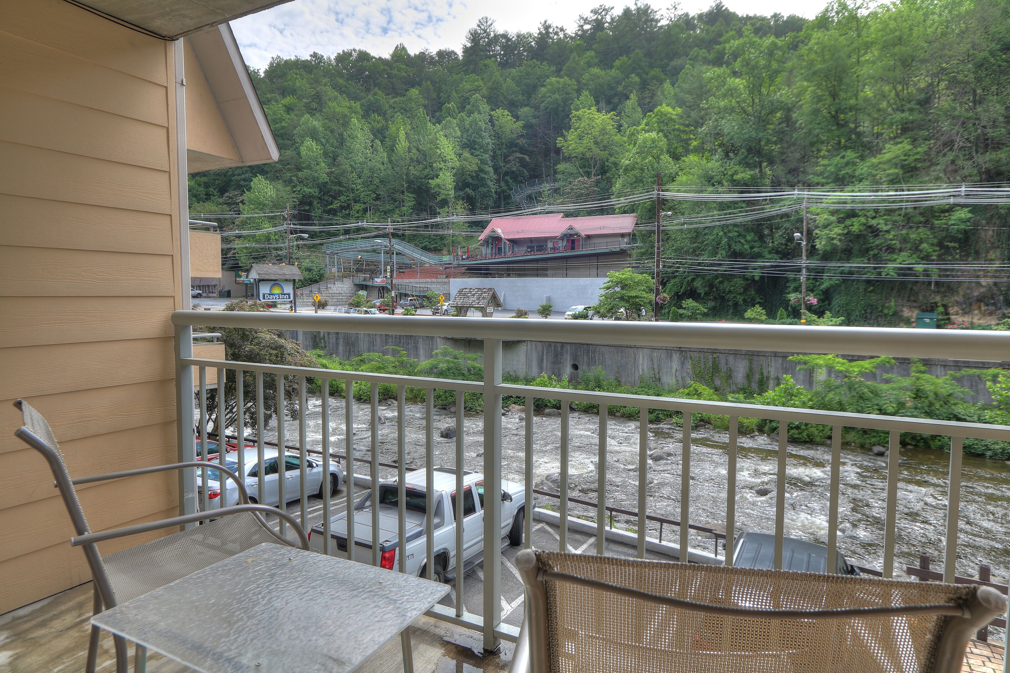 A balcony with metal railings, a table, and a chair. In the background, there is a river, a building with a red roof, and a forested hillside.