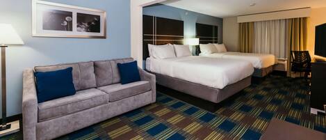 Una habitación de hotel con sofá, dos camas, alfombra estampada, un cuadro en la pared y televisión.