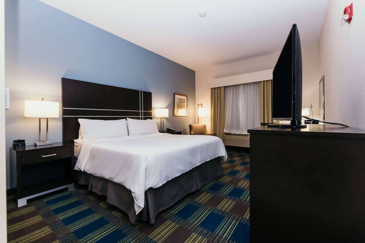 Een hotelkamer met een groot bed, twee nachtkastjes met lampen, een flatscreen televisie, een bureau en een raam met gordijnen.