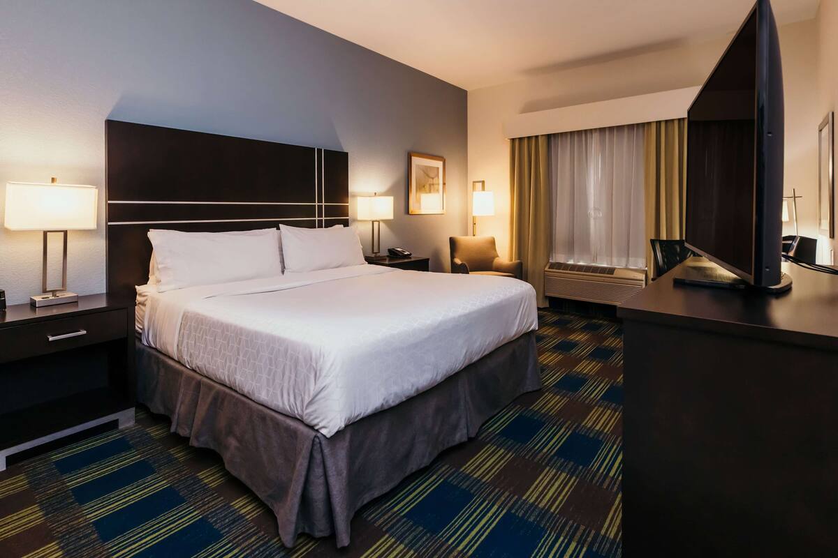 Een hotelkamer met een groot bed, twee nachtkastjes met lampen, een bureau met een flatscreen-tv en een raam met gordijnen.
