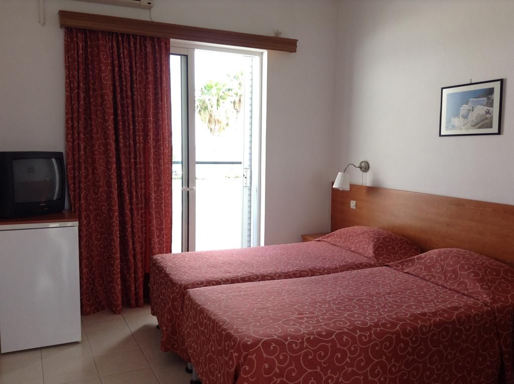 Una habitación con dos camas, un escritorio con televisor, una ventana con cortinas y una lámpara fijada a la pared.