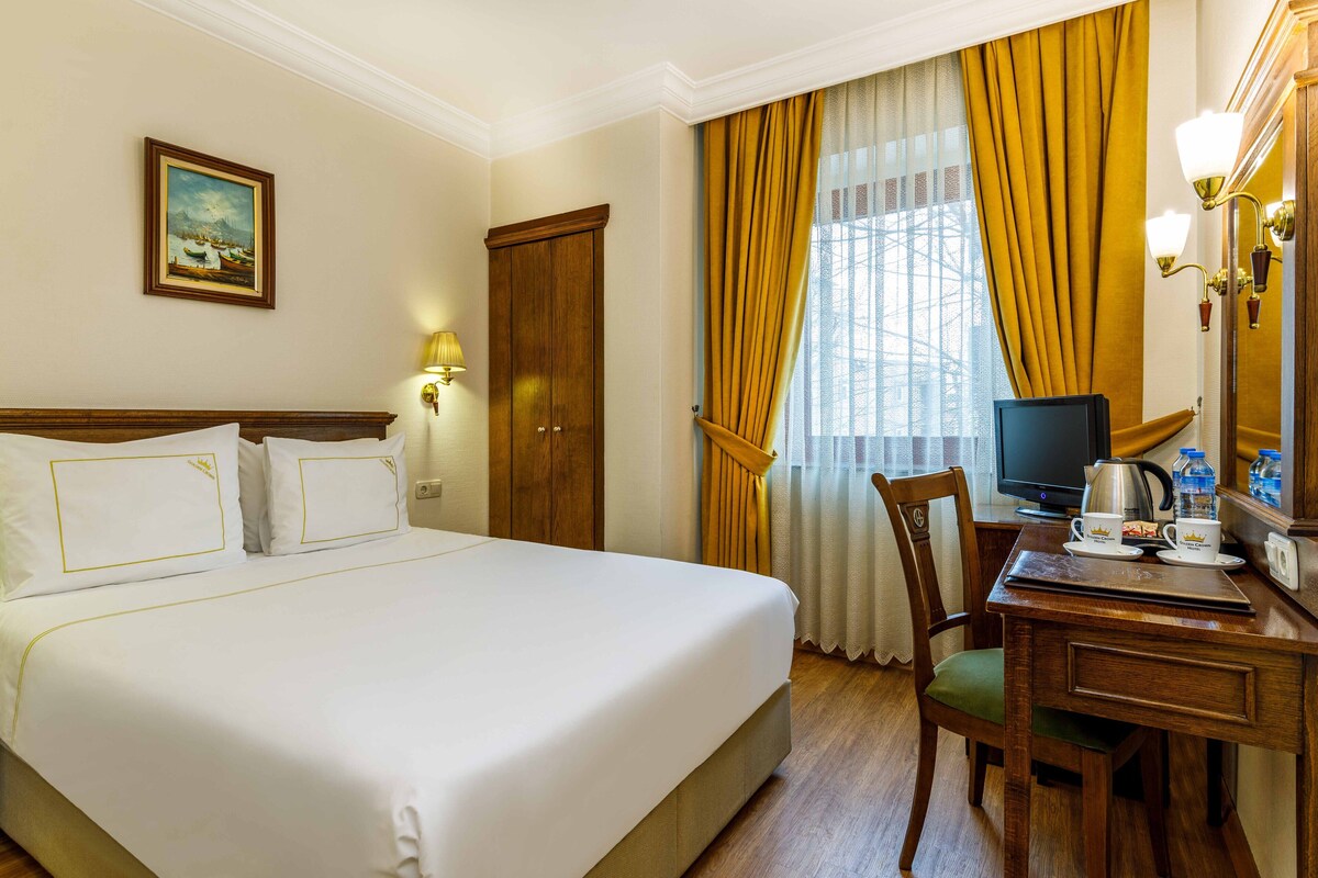 Ein Hotelzimmer mit einem großen Bett, einem Schreibtisch mit Stuhl, einem Fernseher und einem Fenster mit Vorhängen.